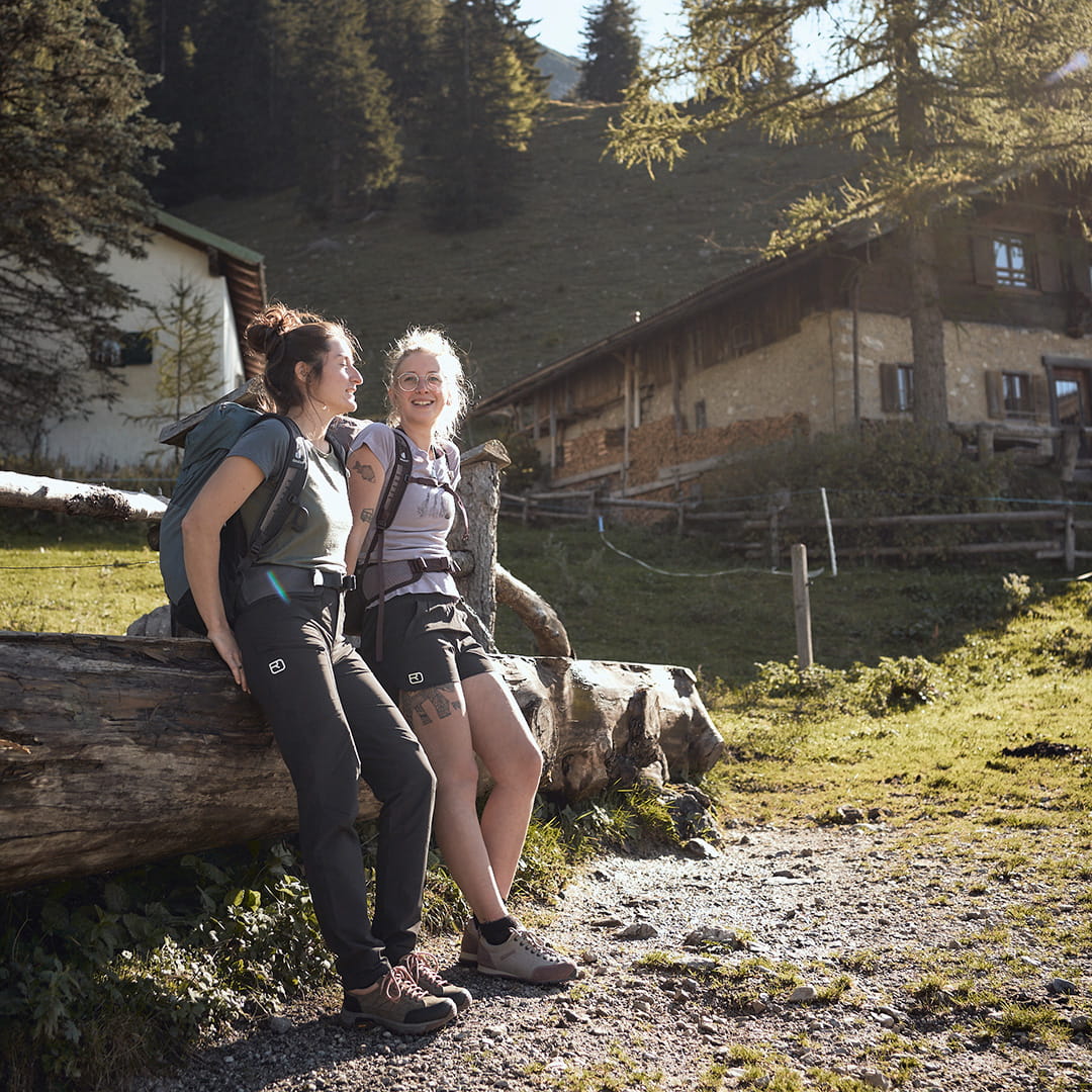 zwei frauen lehnen an einem baumstamm vor einer berghütte und tragen bergschuhe